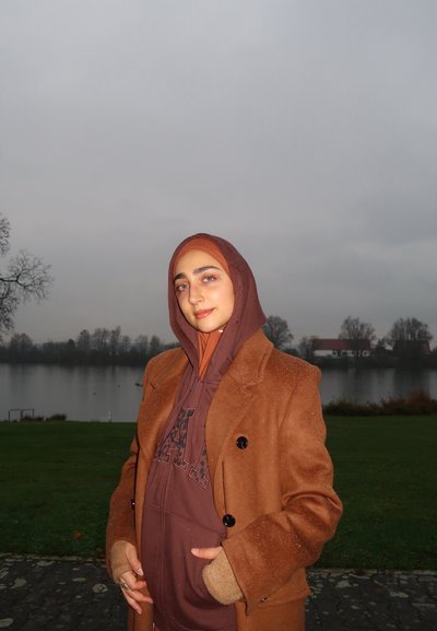 Abrigo marrón de doble botonadura sobre una sudadera con capucha marrón; hiyab a juego; al aire libre junto a un lago; cielo nublado; gotas ligeras en el abrigo.