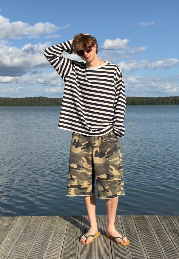 Jeune homme portant des lunettes de soleil, une chemise rayée à manches longues, un short camouflage et des tongs, se tient sur un quai en bois près d'un lac calme.
