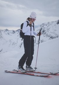 Femme en tenue d'hiver qui fait du ski avec des bâtons sur une pente de montagne enneigée sous un ciel nuageux.