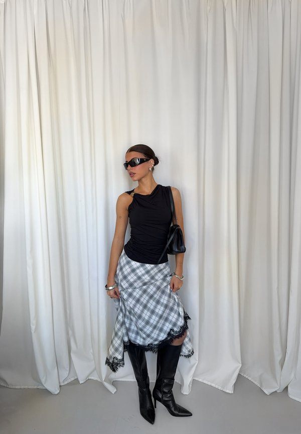Une femme pose devant des rideaux blancs, vêtue d’un haut noir, d’une jupe à carreaux bordée de dentelle, de bottes montantes jusqu’aux genoux, de lunettes de soleil et d’un sac à bandoulière.