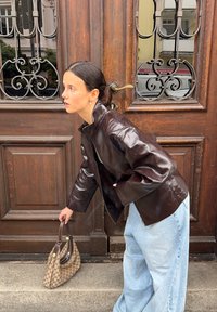 Veste en cuir marron avec un col haut, associée à un jean large bleu clair et un sac à main beige à motifs, devant une porte en bois.