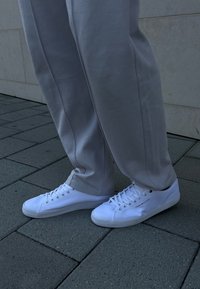 Sneaker bianchi in tela con suola piatta, punta rotonda e stringhe tradizionali. Abbinati a pantaloni a sigaretta grigio chiaro. Pavimentazione grigia testurizzata sullo sfondo.