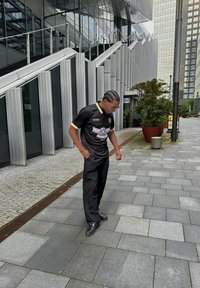 Μαύρο πόλο μπλουζάκι με κίτρινες λεπτομέρειες και γραφικό σχέδιο, σε συνδυασμό με μαύρο παντελόνι και παπούτσια, που στέκεται σε ένα γκρι λιθόστρωτο μονοπάτι κοντά σε σύγχρονη αρχιτεκτονική.