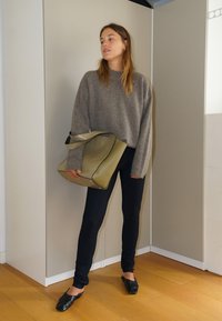 Maglione grigio oversize, leggings neri e scarpe basse nere. Tiene una borsa verde con un design strutturato e una tracolla curva su uno sfondo neutro.