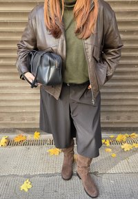 Hnedá kožená bunda cez zelený rolák, doplnená o čierne široké nohavice, hnedé semišové čižmy po kolená a malú čiernu tašku.