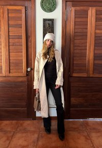 Cappotto lungo beige, top aderente nero, pantaloni a zampa neri e stivali con tacco neri. Berretto lavorato a maglia beige, tiene una borsa a tracolla marrone a fantasia. Sfondo in legno.