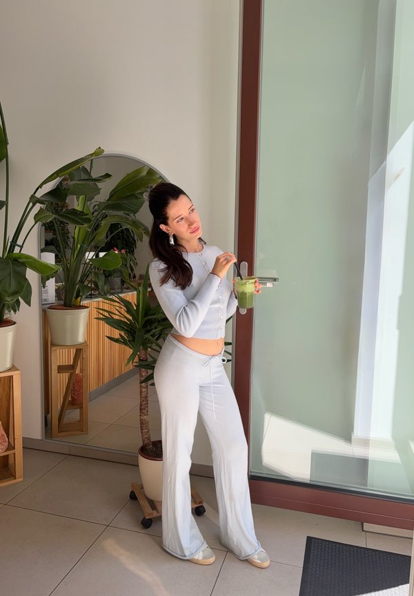 Mujer con ropa cómoda clara sosteniendo una pajita para batido verde junto a una puerta de cristal con plantas en macetas y un gran espejo en una habitación luminosa.