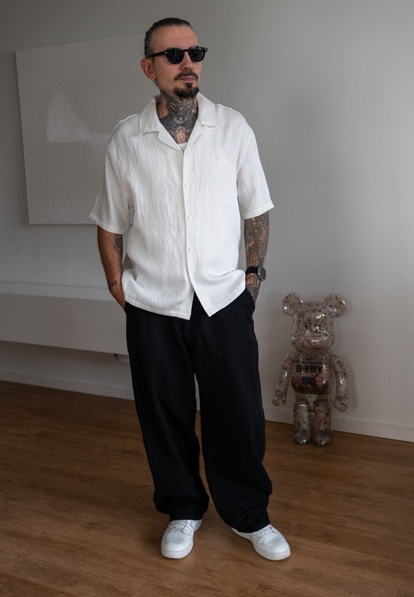 Homme avec tatouages, lunettes de soleil, chemise blanche à manches courtes, pantalon noir et chaussures blanches debout sur un sol en bois près d'une figure transparente en forme d'ours.