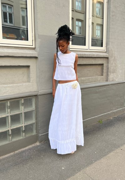 Conjunto blanco de dos piezas que incluye un top corto sin mangas y una falda larga con un dobladillo con volantes, adornado con una flor de tela en la cinturilla.
