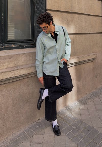 Joven con gafas de sol, camisa verde claro, pantalones negros, calcetines blancos y mocasines negros, parado en una pierna cerca de un edificio de piedra.