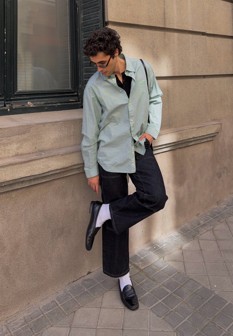 Jeune homme portant des lunettes de soleil, une chemise vert clair, un pantalon noir, des chaussettes blanches et des mocassins noirs, se tenant sur une jambe près d'un bâtiment en pierre.