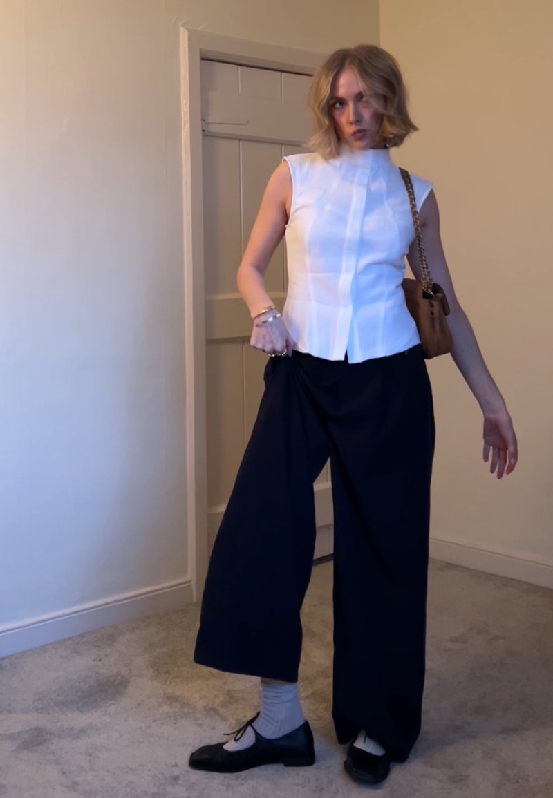 Femme aux cheveux blonds courts portant un haut blanc sans manches, un pantalon large noir, des chaussettes grises, des chaussures noires et un sac à épaule marron à l'intérieur.