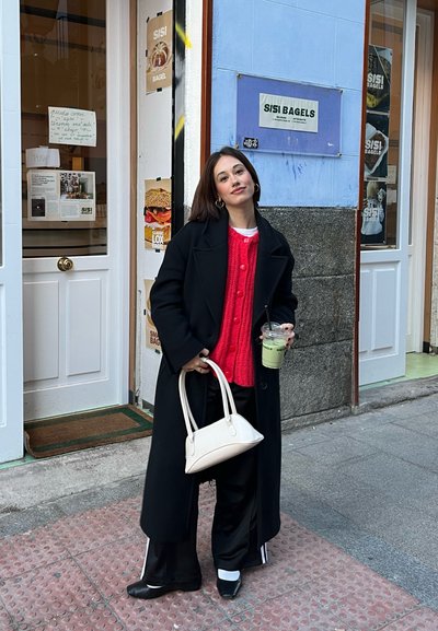 Abrigo largo negro, suéter de punto rojo, pantalones anchos negros con rayas blancas, bolso blanco, sosteniendo una bebida verde fuera de una tienda de bagels.