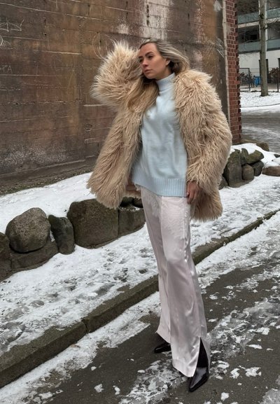 Mujer con abrigo de piel sintética beige, suéter de cuello alto azul claro, pantalones de satén blancos y botas negras, de pie en una acera nevada junto a una pared de ladrillos.