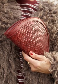 Pochette en cuir tressé rouge avec un dessus incurvé et des bords lisses. La texture présente un motif en forme de panier, tenue par une main aux ongles vernis.