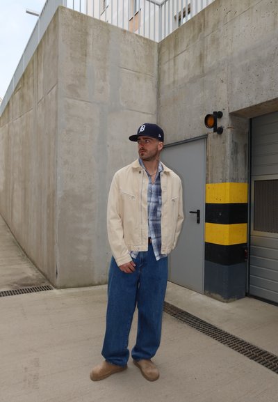 Hombre con chaqueta beige, camisa de cuadros, vaqueros azules y gorra de béisbol azul marino está junto a una pared de hormigón y una puerta industrial con franjas negras y amarillas.