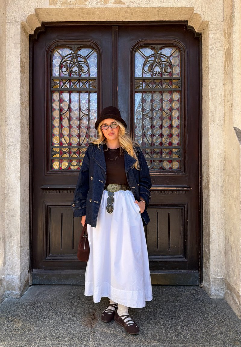 Žena so svetlými vlasmi a okuliarmi nosí čierny klobúk, tmavú bundu, čierny top, bielu sukňu a hnedé topánky, stojí pred ozdobnými tmavými drevenými dverami.