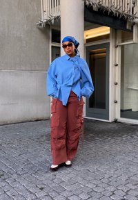 Camicia oversize azzurro chiaro con nodo sul davanti, abbinata a pantaloni cargo color ruggine con ampie tasche e scarpe bianche.