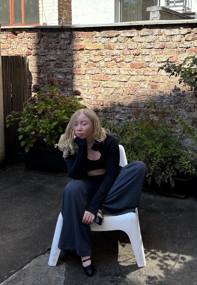 Una persona se sienta en una silla de plástico blanca, vistiendo una camiseta corta de manga larga negra y pantalones grises holgados, con cabello trenzado y zapatos negros.