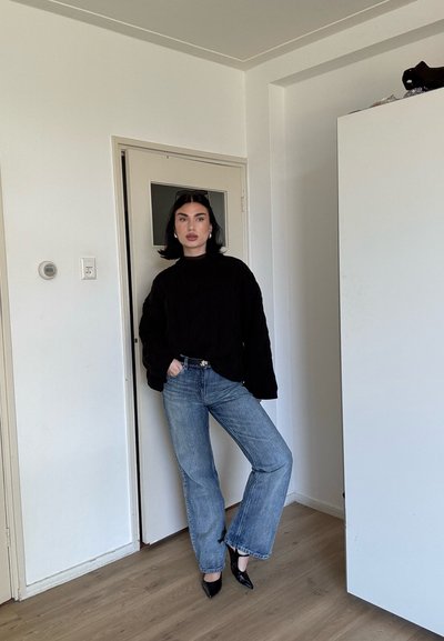 Mujer con cabello negro que viste un suéter negro, jeans azules y tacones negros, de pie en el interior contra una pared y puerta blancas.