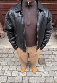 Personne portant une veste en cuir noire par-dessus un pull zippé marron, un pantalon beige et des chaussures marron clair, debout sur un pavé en pierre.