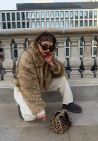 Mākslīgās kažokādas jaka ar brūnām un melnām svītrām, krēmkrāsas bikses, melnās saulesbrilles un neliela leopardu raksta soma, kas atpūšas uz akmens virsmas.