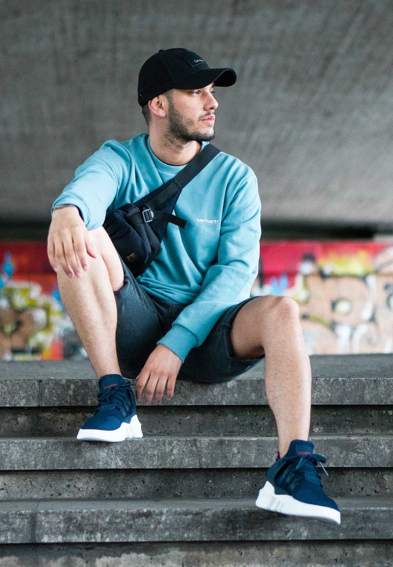 Marineblaue Sport-Sneaker mit weißen Sohlen, hellblauem Sweatshirt, schwarzer Umhängetasche und grauen Shorts, die auf Steinstufen sitzen.