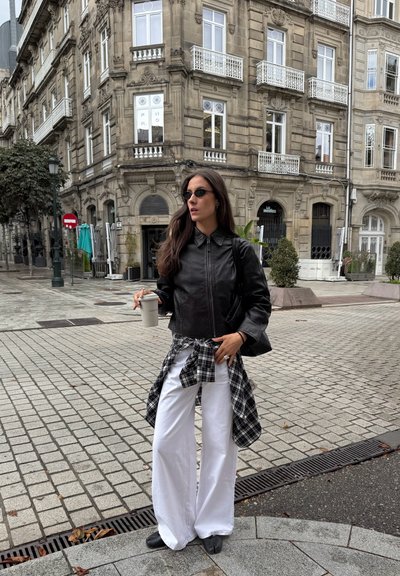 Chaqueta de cuero negra con cuello, combinada con pantalones anchos blancos. Camisa de cuadros en blanco y negro atada a la cintura, sosteniendo una taza de café.