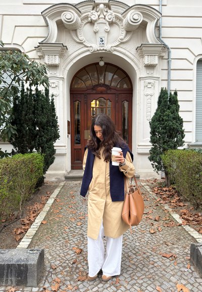 Mujer con cabello largo y oscuro, vestida con abrigo beige y pantalones blancos, de pie en un camino cubierto de hojas, sosteniendo una taza de café y un bolso marrón frente a una puerta ornamentada.