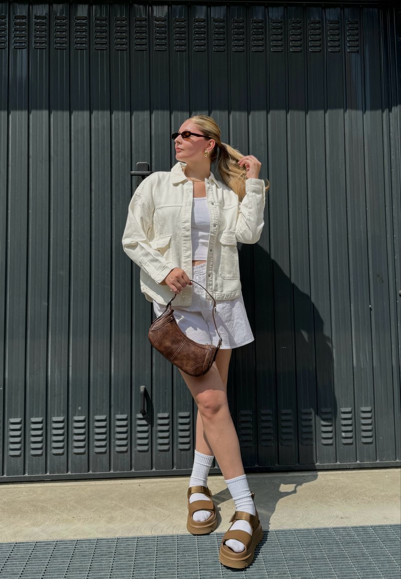 Μια γυναίκα φοράει μια ανοιχτή μπεζ oversized ζακέτα πάνω από ένα λευκό φόρεμα, συνδυασμένη με καφέ πλατφόρμες και μια μικρή καφέ τσάντα.