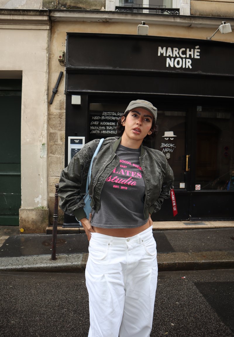 Femme portant une veste grise courte, un t-shirt graphique, un pantalon blanc et une casquette, debout dans une rue mouillée devant une boutique noire avec le panneau « Marché Noir ».