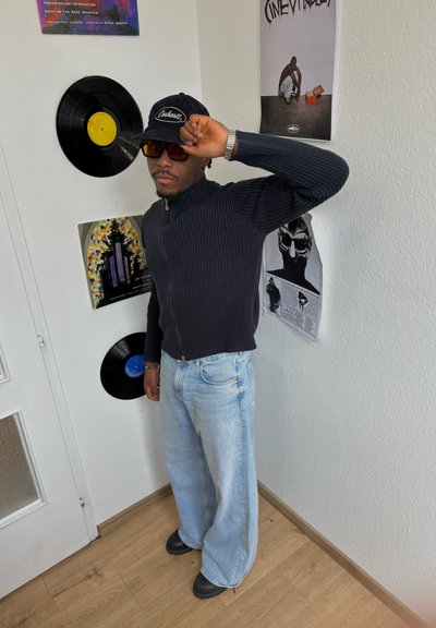Persona que lleva un suéter oscuro de canalé con cierre, unos vaqueros anchos de color azul claro y una gorra negra; gafas de sol; discos de vinilo en la pared.