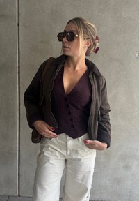 Giacca bomber marrone sopra un top a coste viola scuro con bottoni, abbinata a pantaloni larghi di colore avorio. Grandi occhiali da sole quadrati e una borsa marrone.