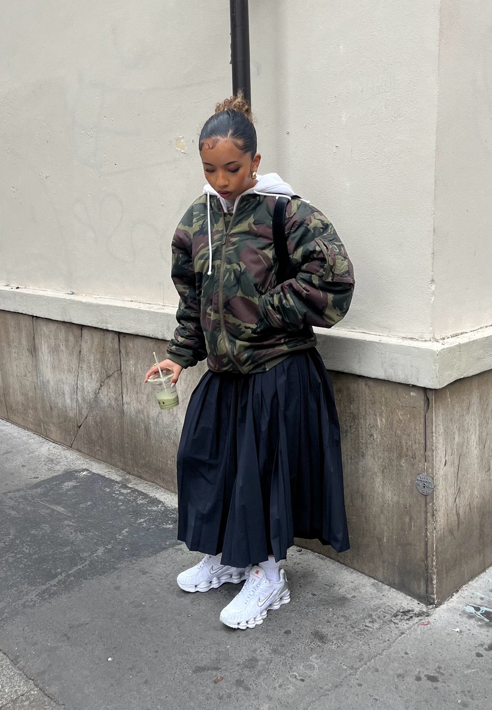 Jeune personne portant une veste camouflage et une jupe plissée noire, tenant une boisson verte, debout à un coin de rue en ville.