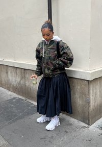 Jeune personne portant une veste camouflage et une jupe plissée noire, tenant une boisson verte, debout à un coin de rue en ville.