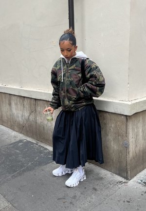 Joven con chaqueta de camuflaje y falda plisada negra, sosteniendo una bebida verde, de pie en una esquina de la calle de la ciudad.
