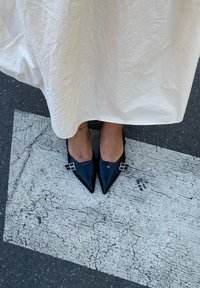 Sapatilhas pretas de verniz com bico apontado e dois fivelas prateadas em cada sapato, usadas com uma saia branca de tecido texturizado. Pés posicionados sobre o pavimento.