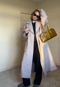 Femme portant un long manteau gris clair, des lunettes de soleil noires, un pantalon noir et des chaussures grises, tenant un grand sac à main vert olive à l'intérieur.