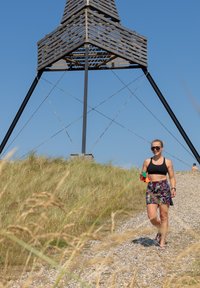 Uma mulher com uma blusa preta e calções floridos caminha por um caminho de cascalho. Uma estrutura de madeira com um design angular ergue-se ao fundo.