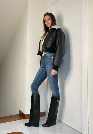 Chaqueta de cuero negra corta con cuello de borreguito, combinada con vaqueros azules ajustados y botas negras altas de diseño ancho y tacón bajo.