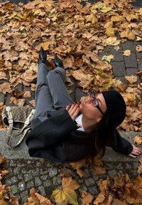 Šedé společenské kalhoty a černý svetr jsou kombinovány se zelenou čepicí a černými špičatými botami. Texturovaná kabelka je na zemi mezi opadaným listím.