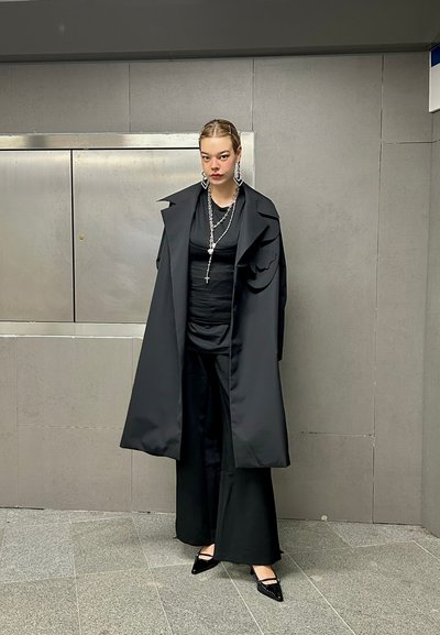Abrigo negro largo con solapas anchas, superpuesto a una blusa negra y pantalones de pierna ancha. Los accesorios incluyen aretes de plata grandes y cadenas.