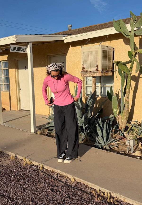 Femeie cu cămașă roz cu mâneci lungi și pantaloni negri stând pe trotuar în fața unei clădiri galbene de spălătorie cu plante deșertice în apropiere.