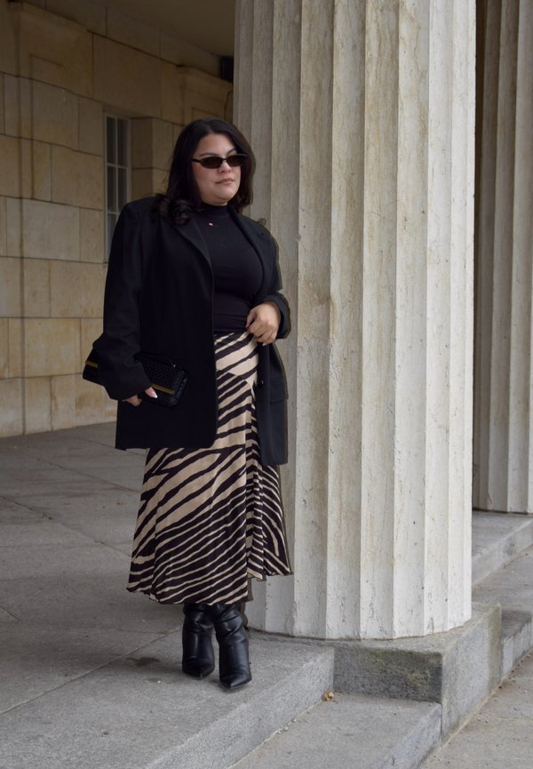 Una mujer con gafas de sol, abrigo negro, falda de rayas y botas está de pie junto a una columna de piedra, en el exterior de un edificio.
