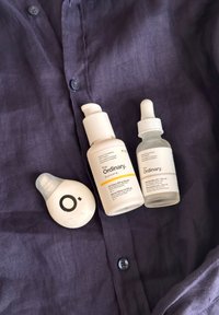 Tres productos de cuidado de la piel sobre una camisa morada oscura: dos botellas blancas con dispensadores y goteros, además de un recipiente redondo blanco con un logo negro.