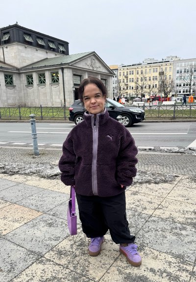 Chaqueta de forro polar morada con cuello alto, pantalones holgados negros y zapatos morados. Llevando un pequeño bolso morado, de pie sobre un pavimento de piedra.