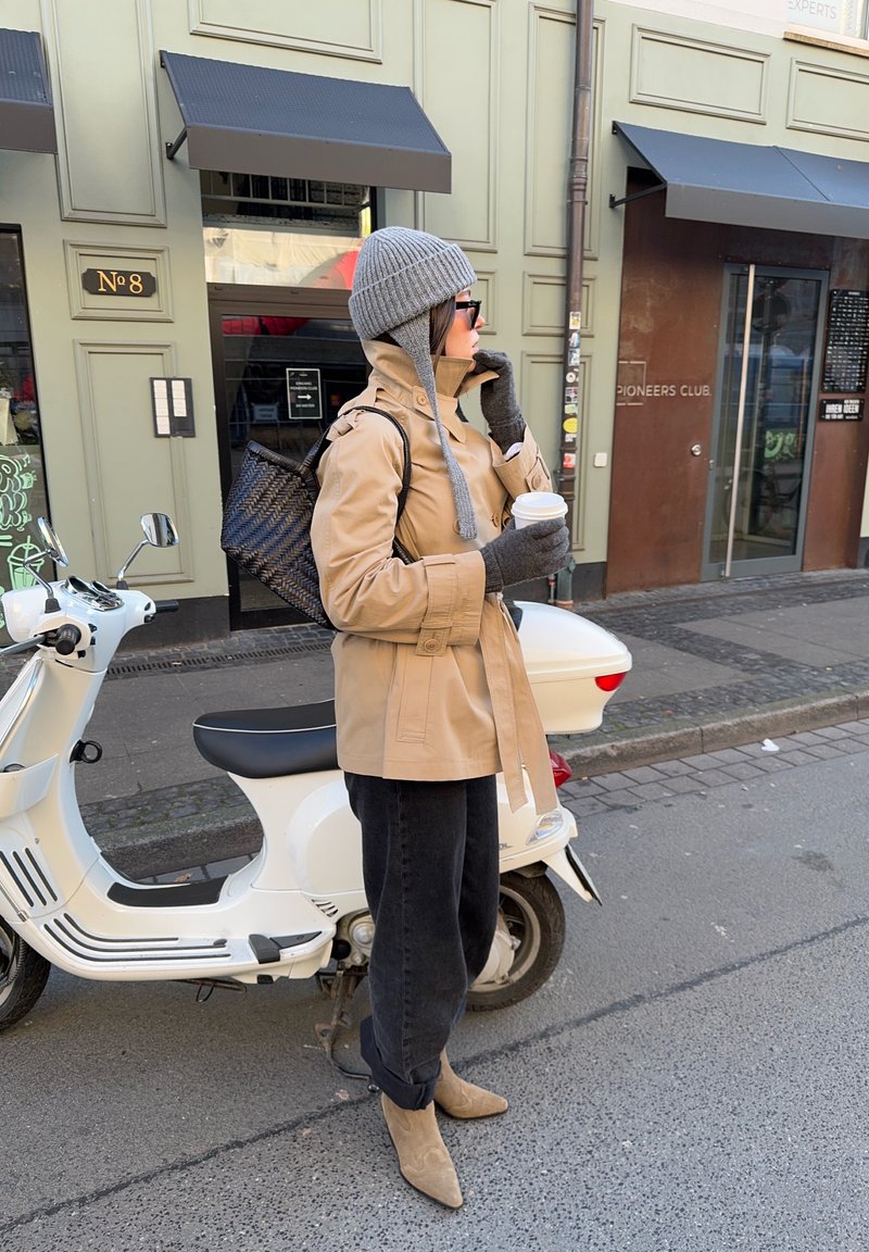 Veste légère beige, bonnet en maille gris, gants gris. Pantalon noir, bottes couleur sable. Tenant une tasse de café blanche, debout à côté d'un scooter blanc.
