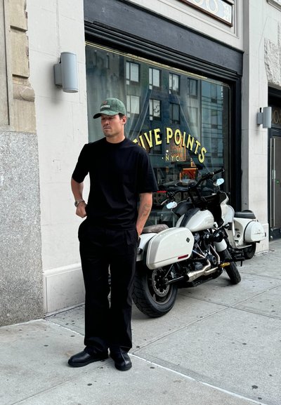 Camisa negra de manga corta y pantalones de textura suave; gorra verde; motocicleta blanca con acentos de cromo aparcada cerca; fondo de una tienda urbana.