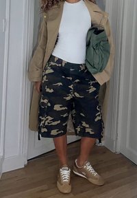 Osoba ubrana w beżowy płaszcz trencz, biały top bez rękawów, moro szorty cargo, beżowe trampki, trzymająca zieloną torbę pod ramieniem.