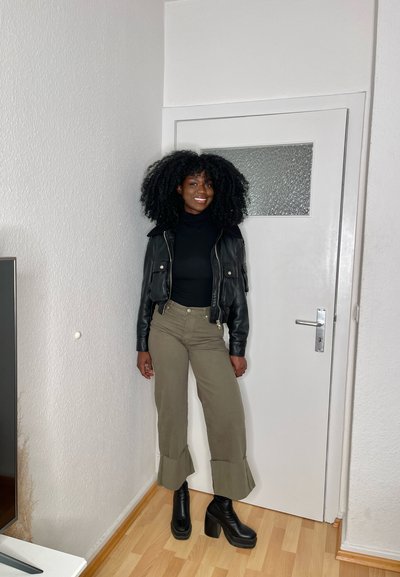 Chaqueta de cuero negra recortada, suéter negro de cuello alto, pantalones verde oliva de pierna ancha con dobladillo acampanado y botas de plataforma negras. Suelo de madera clara, paredes blancas.